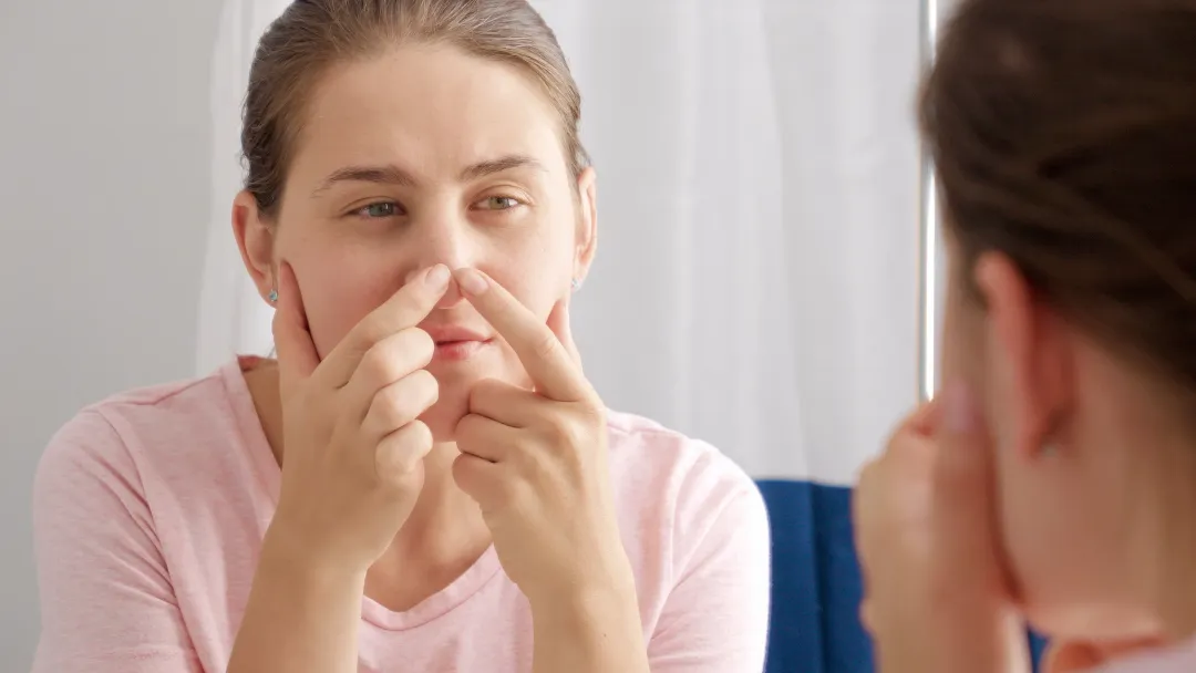 Young woman looking in mirror and squeezing pimples on her nose - How to Get Rid of Blackheads on Nose and Ears - Palm Beach Dermatology Group - PGA Dermatology